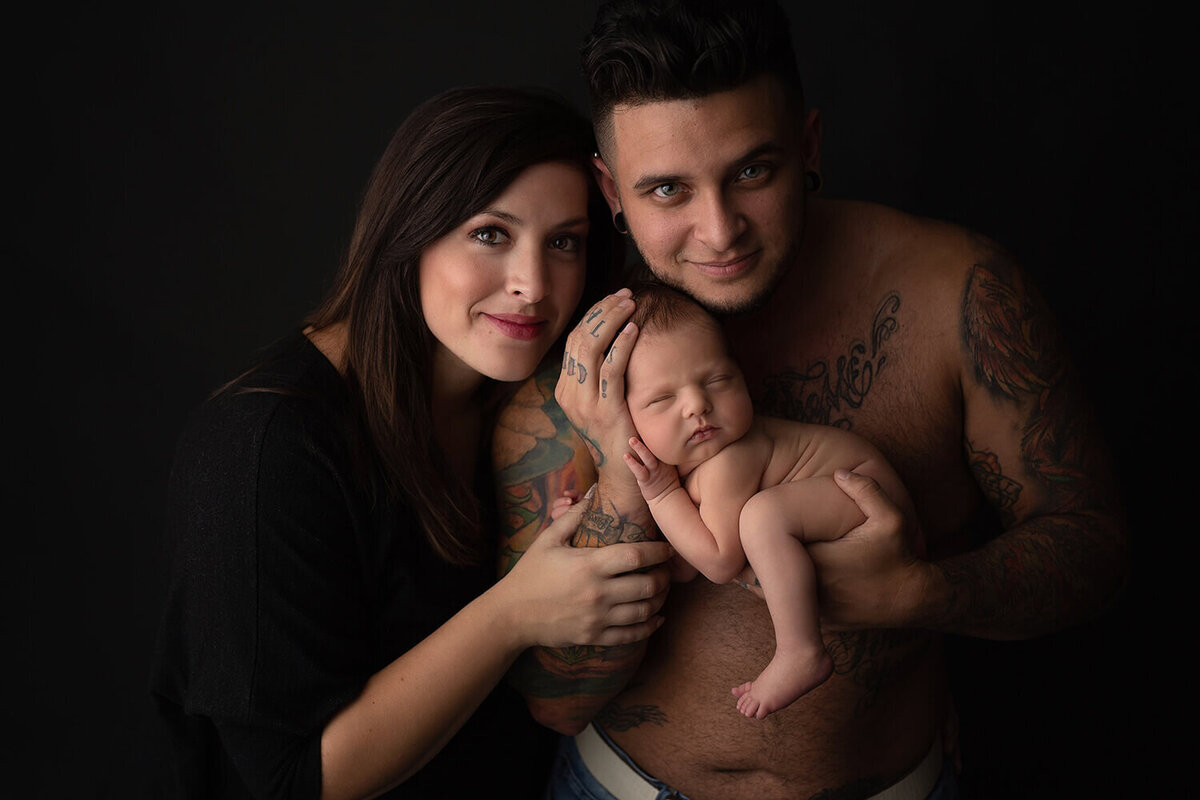 A father with no shirt cradles his sleeping newborn baby with mom leaning on him while standing in a studio posed by a New Orleans Newborn Photographer