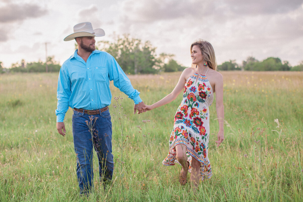 An engaged man wearing a white cowboy hat smiles at his fiancee in a vibrant embroidered floral dress