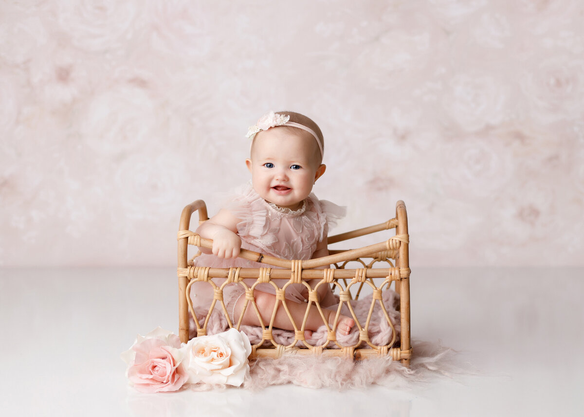 6-month baby photoshoot. Baby girl is wearing a blush organza dress and is sitting in a vintage rattan crib looking over her shoulder and smiling at the camera.  There is a muted dusty rose and white backdrop.