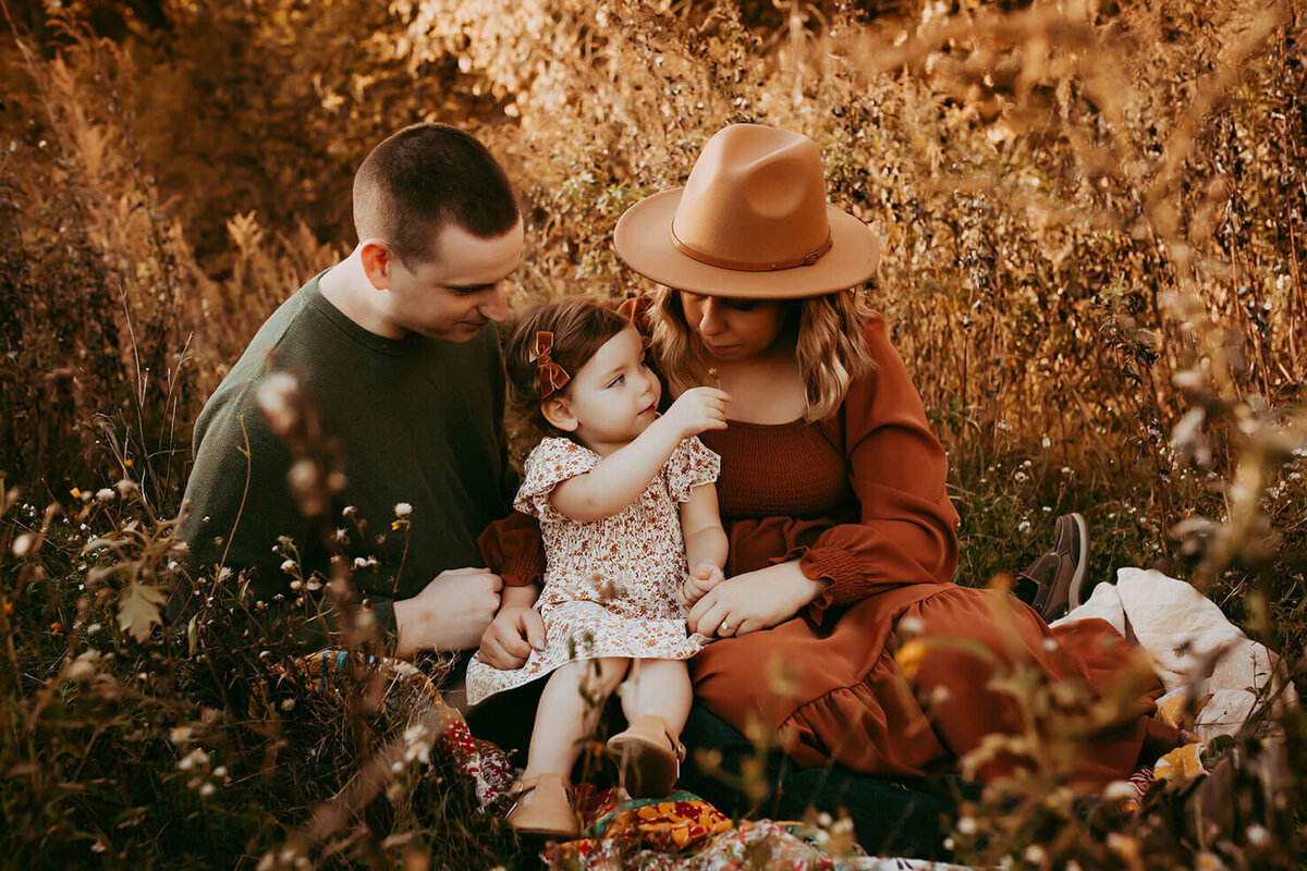 a mom dad and daughter. mom is smelling a flower held by the daughter in a field in rochester ny