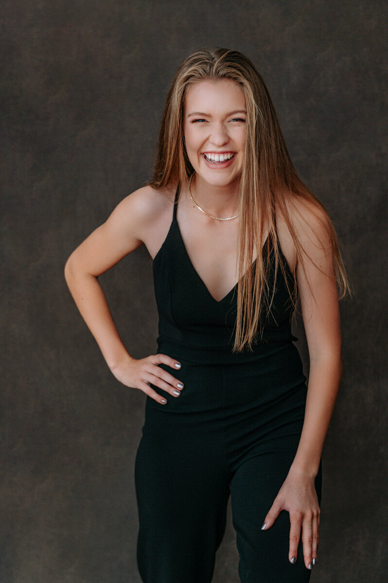 Senior girl in a black jumpsuit, smiling brightly in front of a classic backdrop.