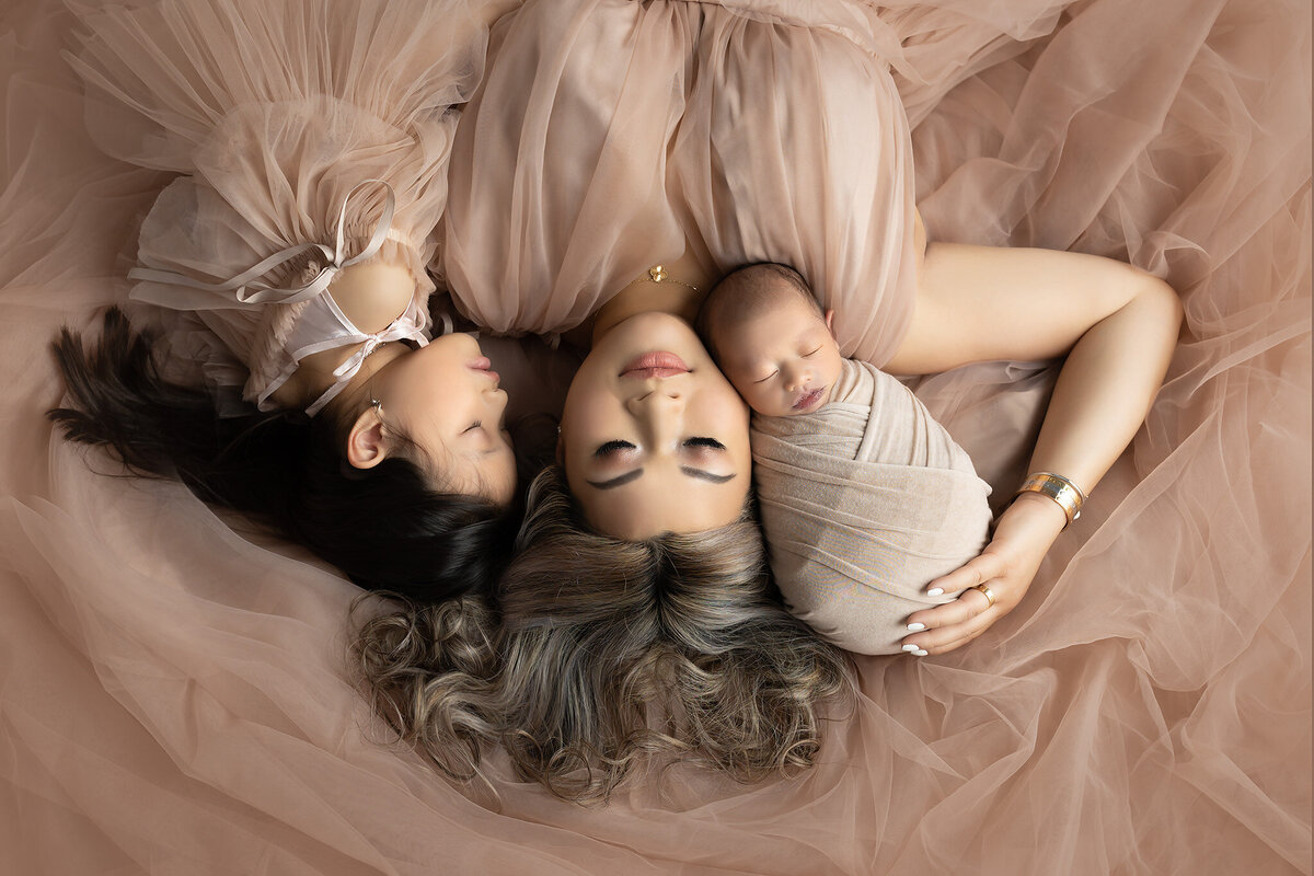 DFW family photo of mom with newborn and young daughter, laying on soft peach tulle in an elegant motherhood portrait
