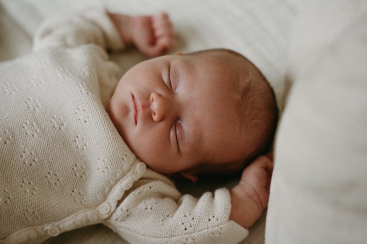 Bébé dort paisiblement, en body tricoté, poing levé au-dessus de sa tête. Shooting photo naissance à domicile par Kamescop.