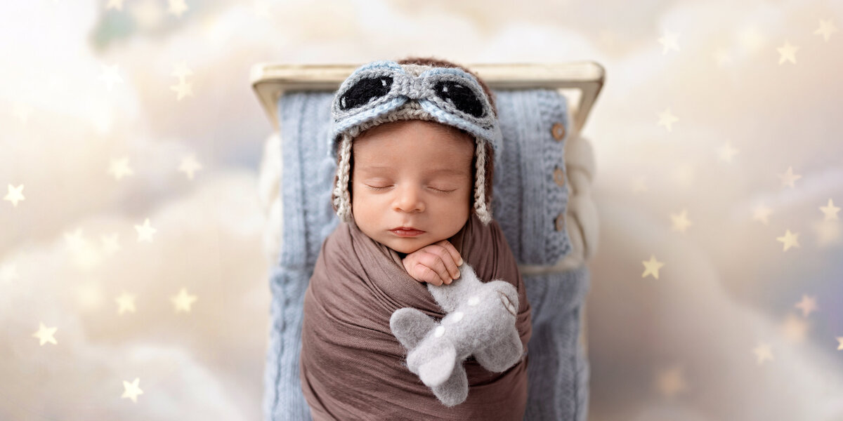 Sleeping newborn baby wrapped in brown, wearing a knit aviator hat and holding a small felt airplane on a starry sky backdrop.