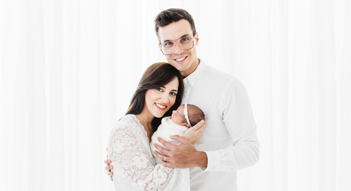 The photograph portrays a heartwarming moment between a mother and father holding their newborn baby. The family members are all dressed in white, standing in an all-white studio. The shot is captured by photographer Bri Sullivan in a pure, clean and classic style, emphasizing the tranquility and innocence of the subjects.