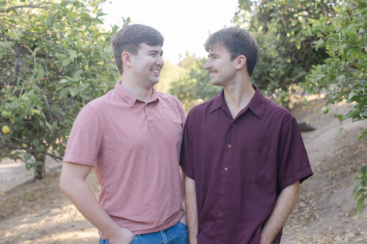 Two brothers wearing different shades of pink with dark hair looking at each other and smiling surrounded by green trees.