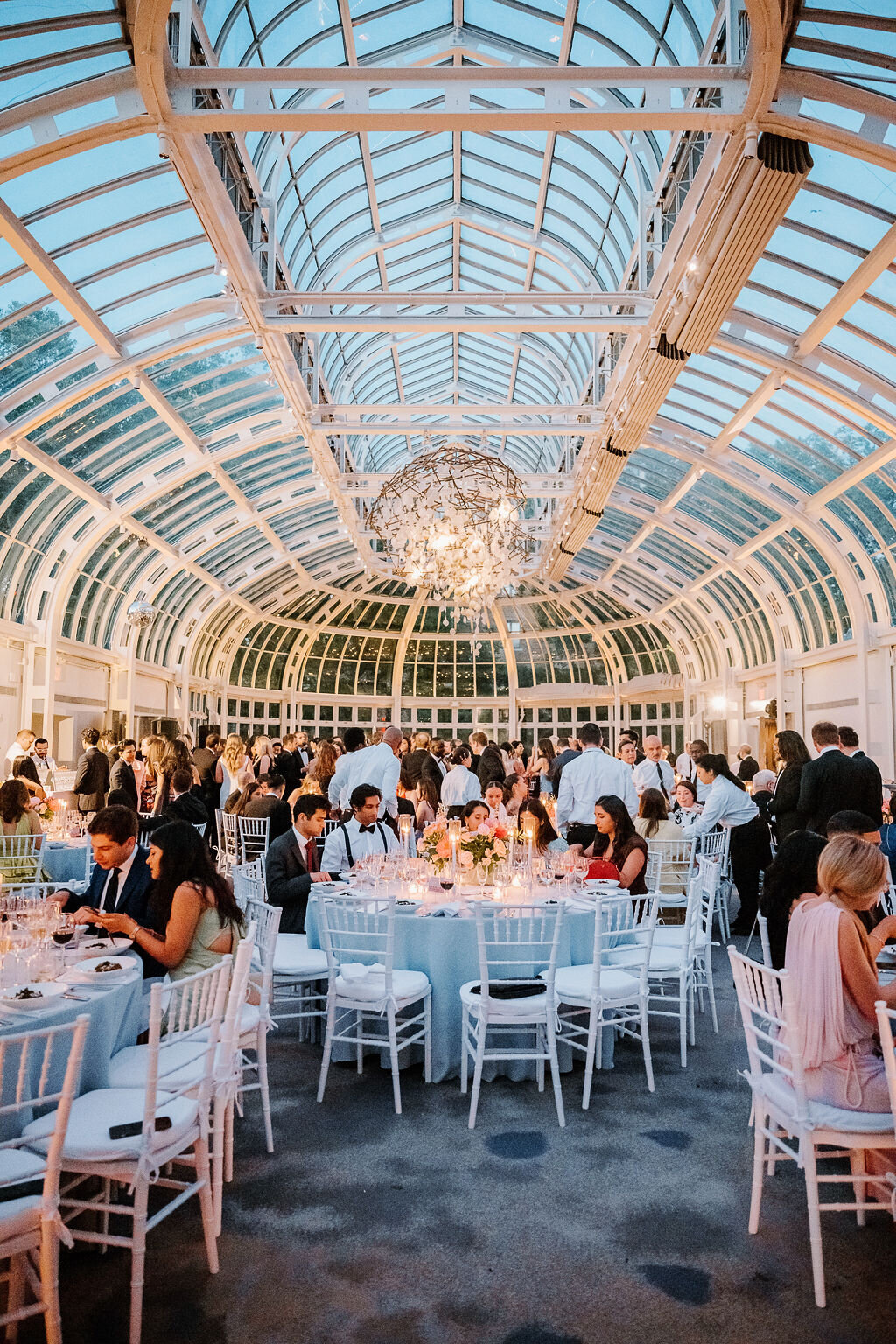 Rhode Island Wedding Photographers | Elegant wedding reception in a glass-ceiling venue with a grand chandelier. Guests in formal attire mingle around beautifully set tables