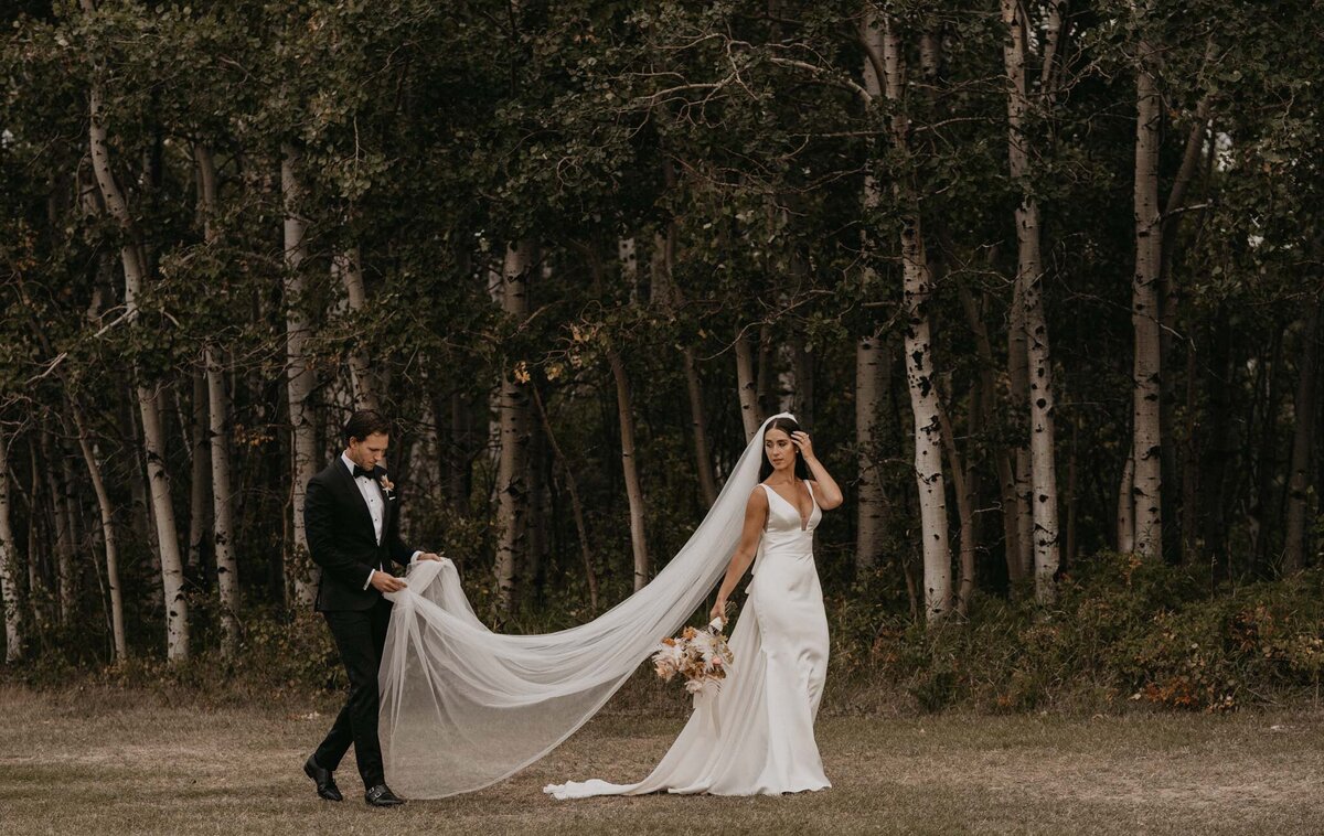 Wedding couple walking, bride wearing a fitted simple satin gown, groom holding long veil, captured by Ash Maclean Photography, romantic elopement and wedding photographer in Red Deer, Alberta. Featured on the Bronte Bride Vendor Guide.