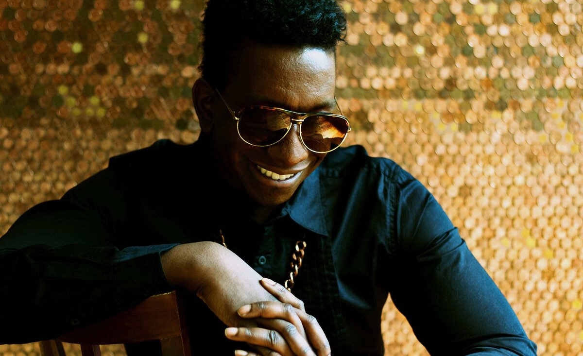 Male musician portrait Thane sitting wearing sunglasses hands clasped gold backdrop looking down smiling