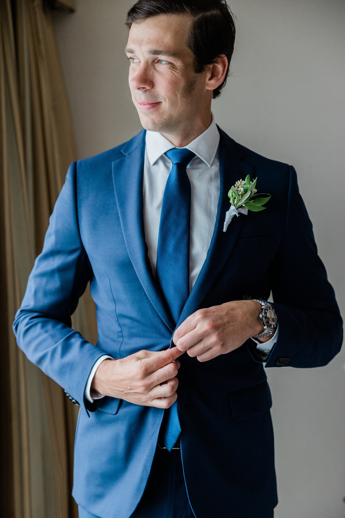 The groom, dressed in a tailored navy suit with a matching tie, buttons his jacket while standing near a window