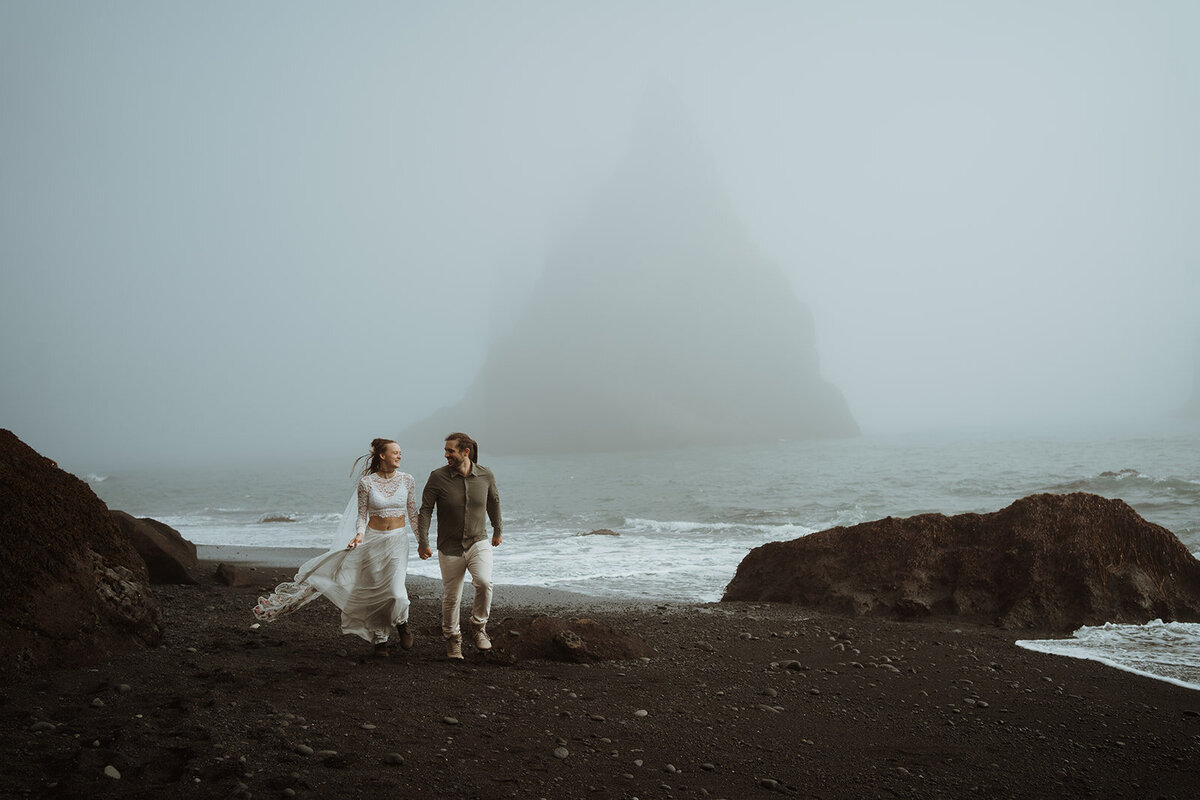 couple is holding hands near the ocean and running towards the camera. They are smiling at each other. There is fog everywhere and waves coming in.