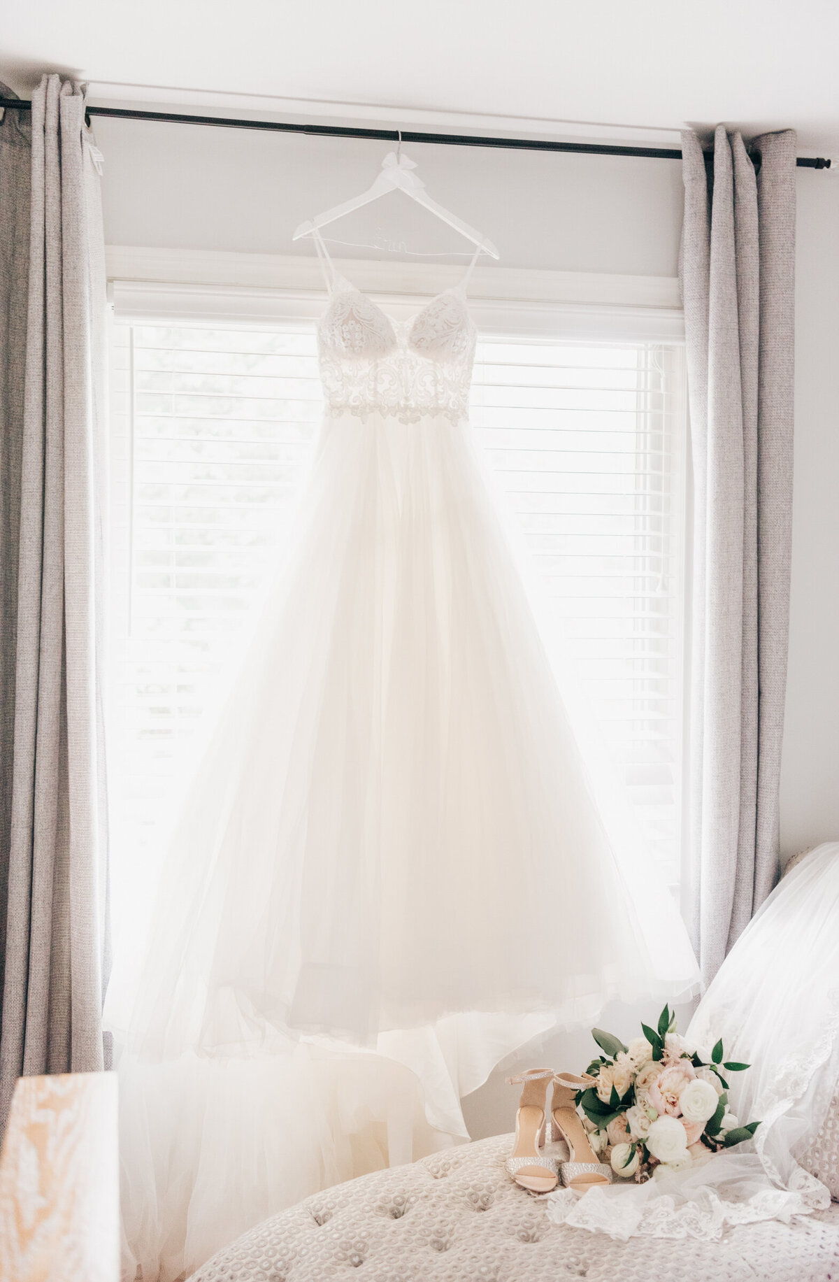 Beautiful white wedding dress hung in window beside white wedding heels and bouquet while getting ready for outdoor wedding