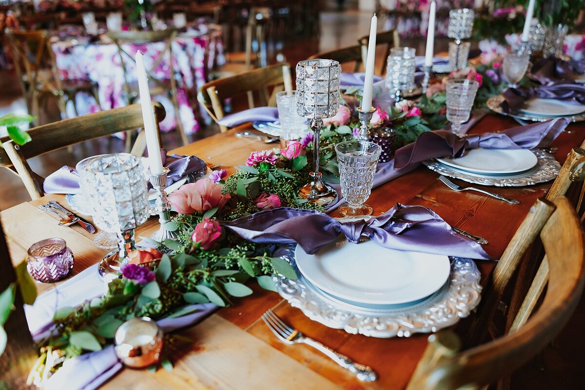 Long farm table with a purple table runner and a long greenery garland with pink roses, blush roses and purple roses. Tall white tape candles in silver candle sticks and low silver votive candles sit on either side of the garland. Ornate silver chargers, white plates and a knotted purple napkin sit at each place setting with a cross back barn wood chair at Steel Magnolia Barn.