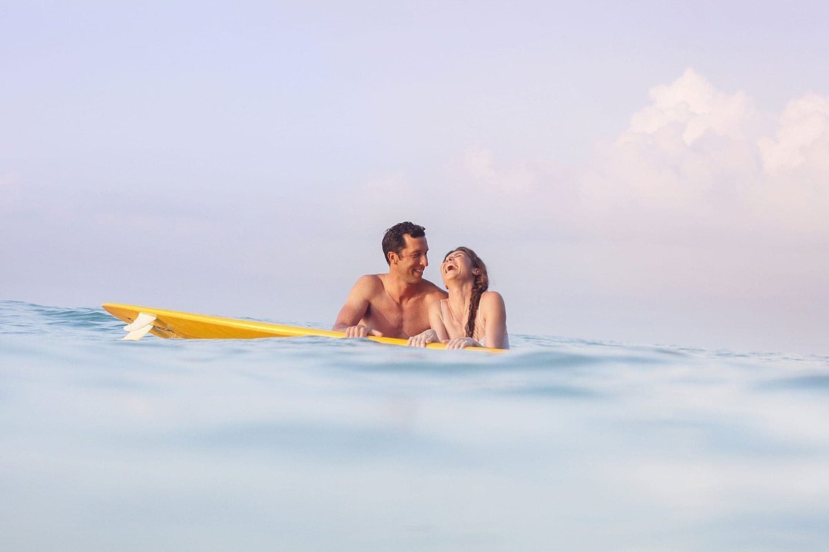 Surfing couple pose on a yellow surfboard as girlfriend looks up at her boyfriend and laughs