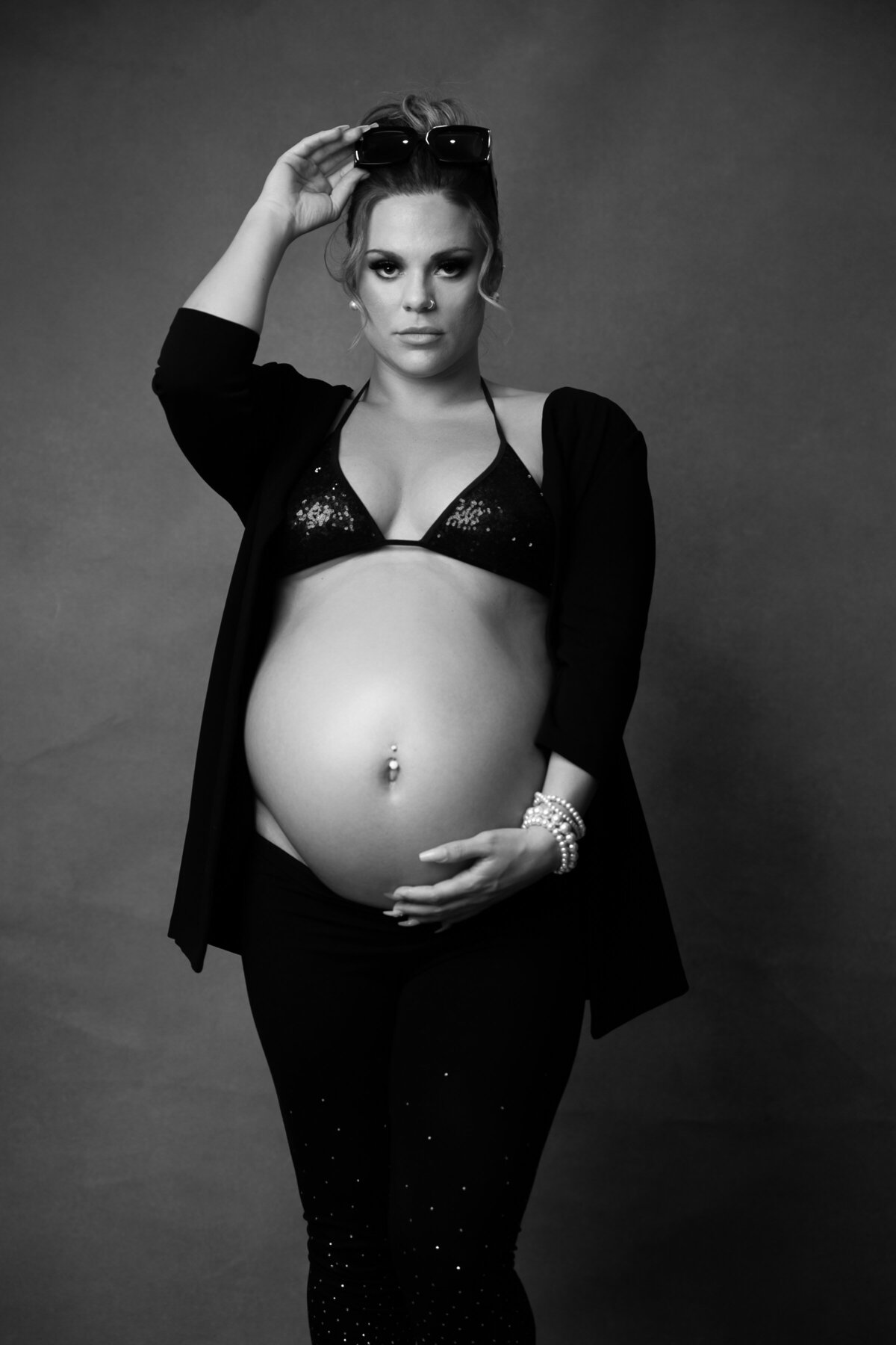 Black and white portrait of a pregnant woman  wearing a sequined bikini top paired with black leggings and an open blazer showing her belly