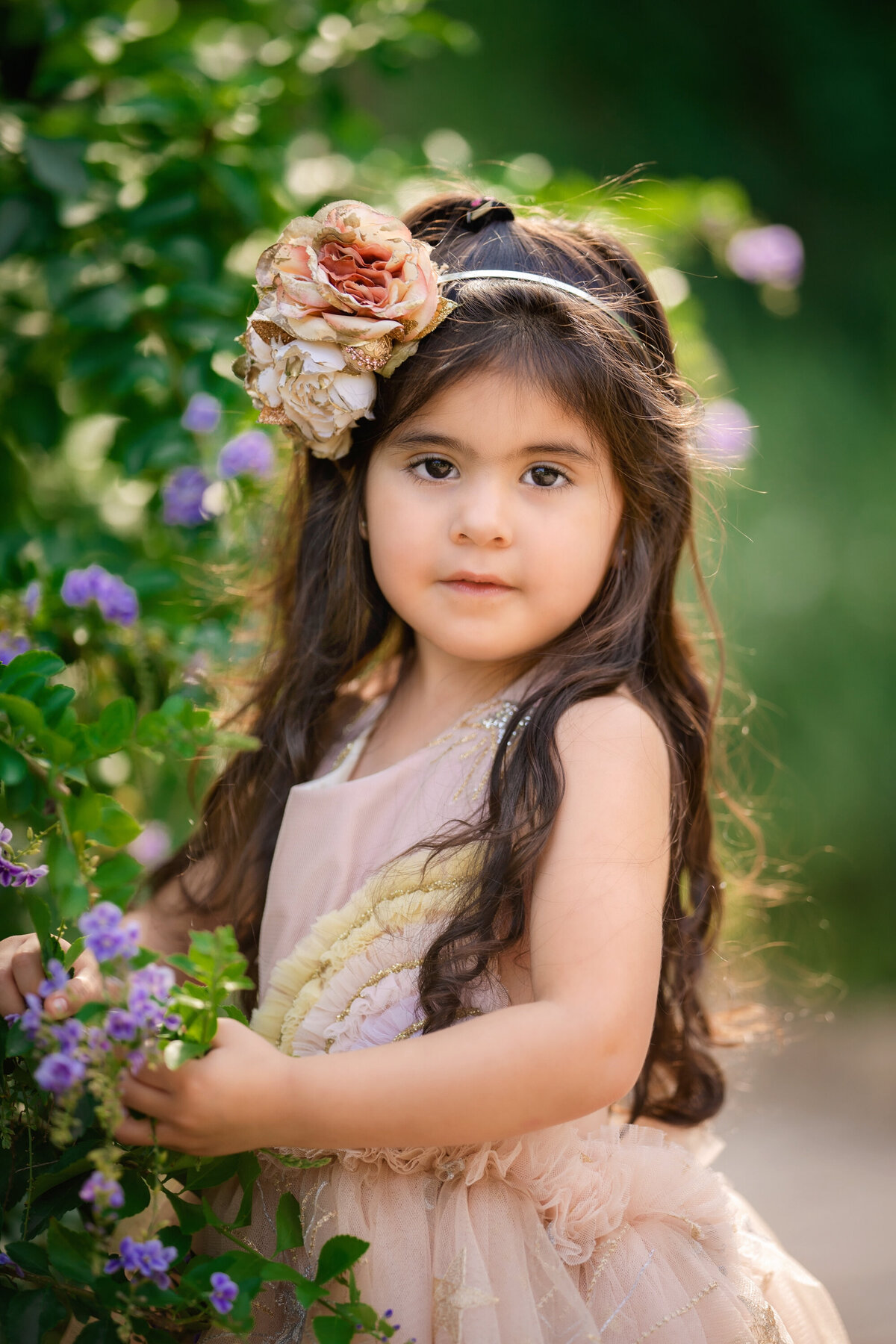 Young girl with very long dark brown hair standing near purple flowers in the Japanese Garden.  She is holding one of the flowers.  She is wearing a pale pink dress with a pastel rainbow from Little Willow Tree.