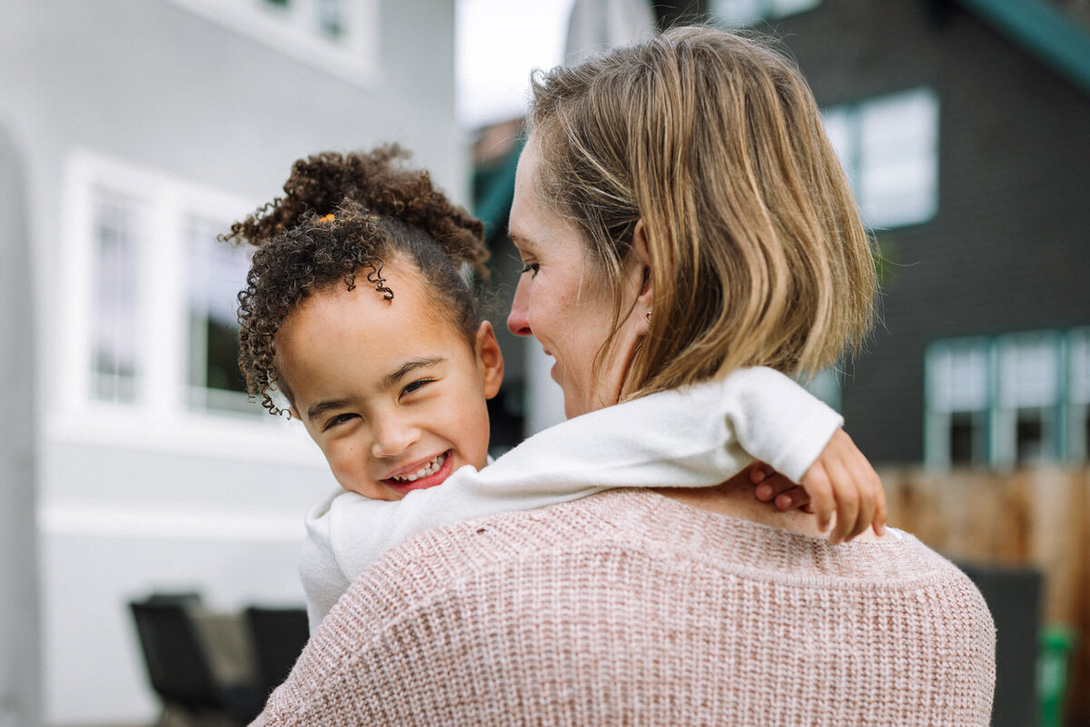 5-year-old girl in moms arms looking over mom's shoulder and laughing as mom looks at her