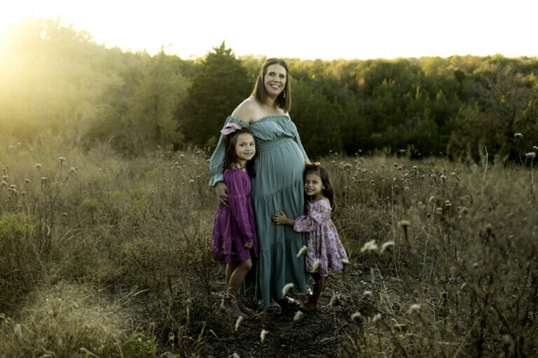 A heartwarming maternity photo of the expectant mom and her two young daughters in the park, with the girls lovingly touching their mom’s baby bump, captured in a joyful moment by Chunky Monkey Photography.