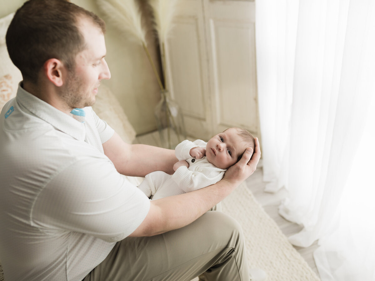 dad holding newborn baby boy in white onesie for studio photoshoot 