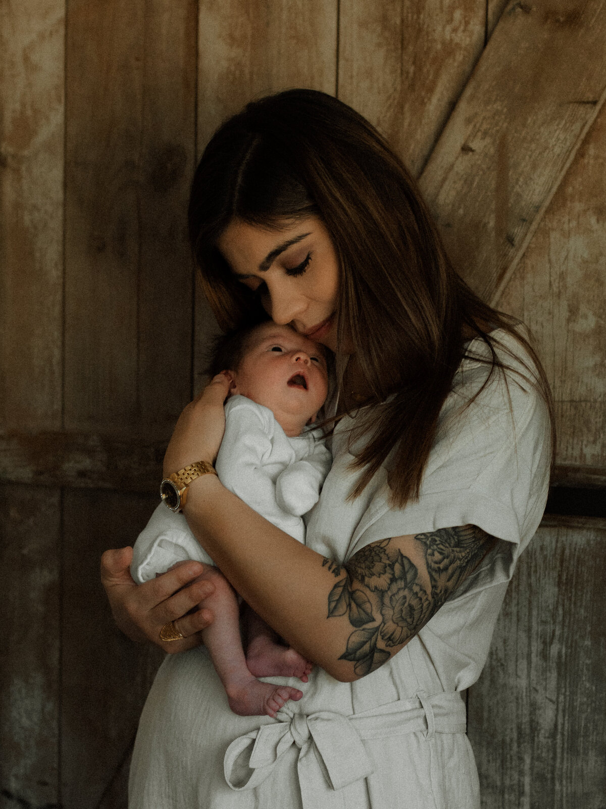 Mother in white romper holds her newborn close against a rustic wooden backdrop during a lifestyle session.