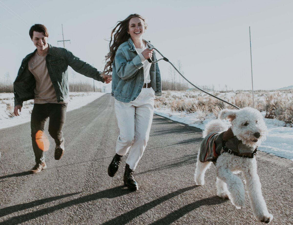 man and woman holding hands and running down a road with their dog on a leash as the couples laughs captured by Idaho Falls wedding photographer