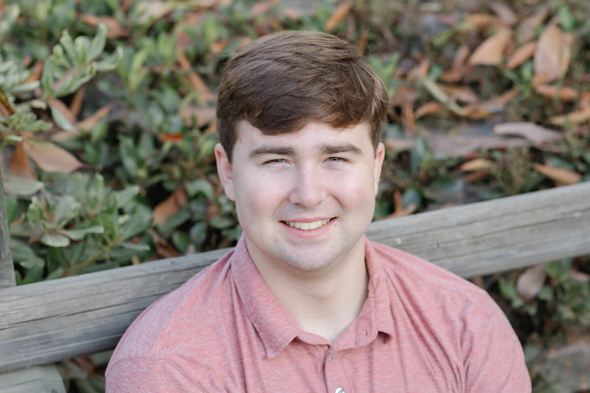 A close-up of a young man with dirty blonde hair smiling and looking up at the camera in a salmon shirt, leaning against a wooden park fence in Tustin.