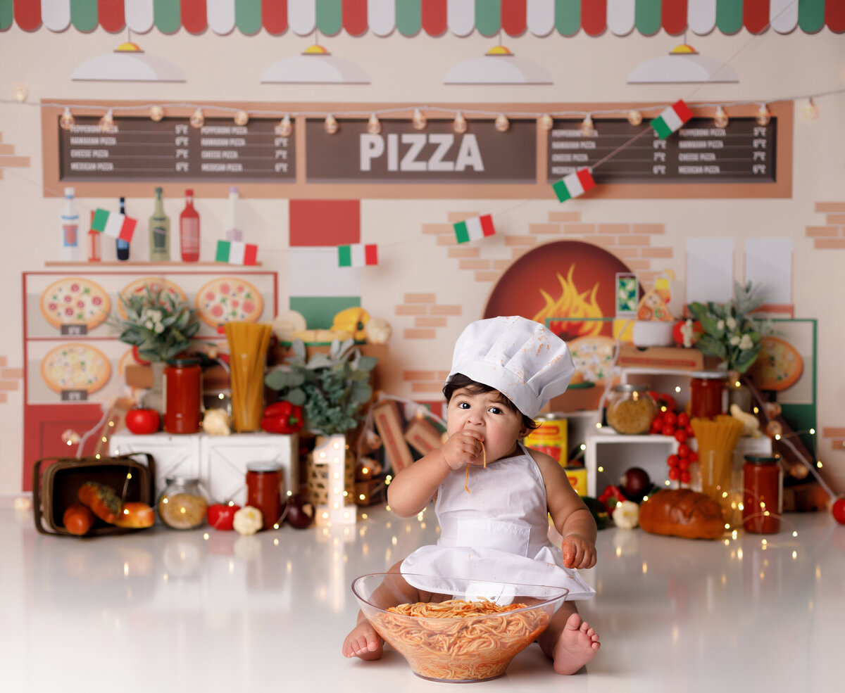 Italian pizza and pasta themed smash in Wellington and West Palm Beach Florida photography studio. Baby boy is wearing a chef inspired outfit. Background is a pizza shop theme with pasta sauce, greens, pasta in jars and various vegetables. Baby boy is eating a bowl of spaghetti.