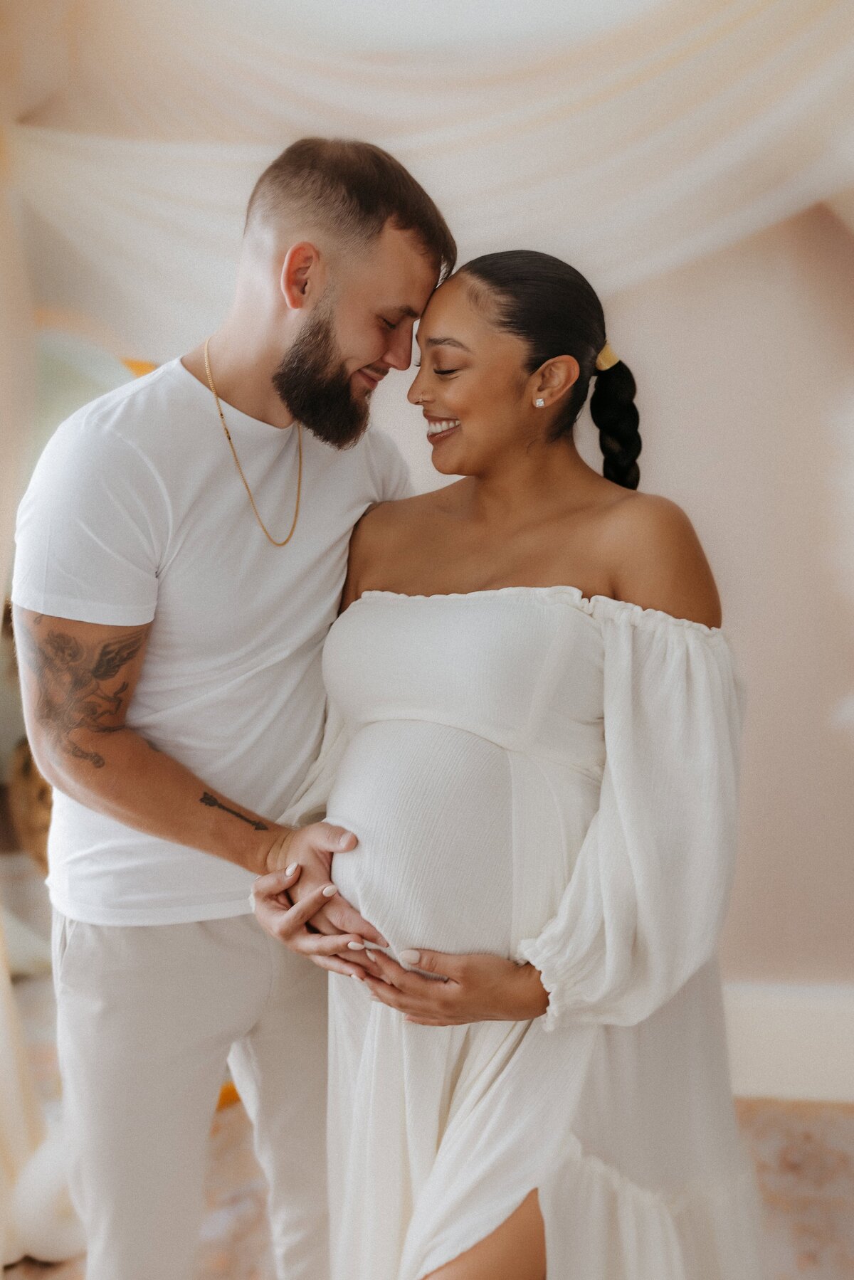 A couple in white outfits stands close, smiling. The woman is pregnant, and both are touching her belly.
