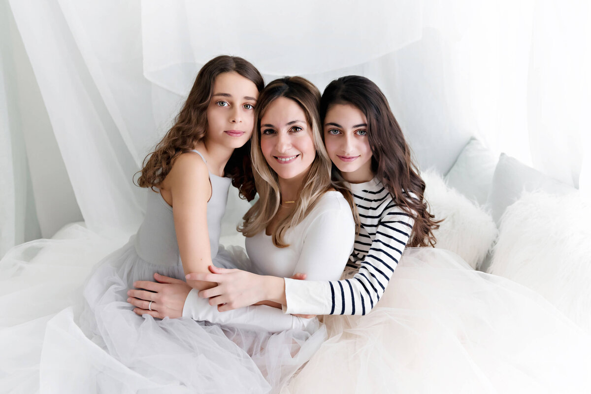 Mom with her teenage daughters sitting around her on a white bed with white draping for her studio family photos
