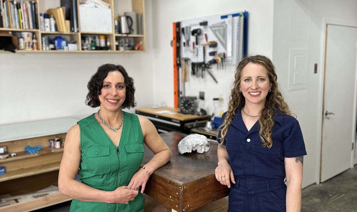 Two printmakers, one in a green sleeveless dress and the other in a navy blue jumpsuit, stand confidently in their printmaking studio surrounded by tools and artwork.