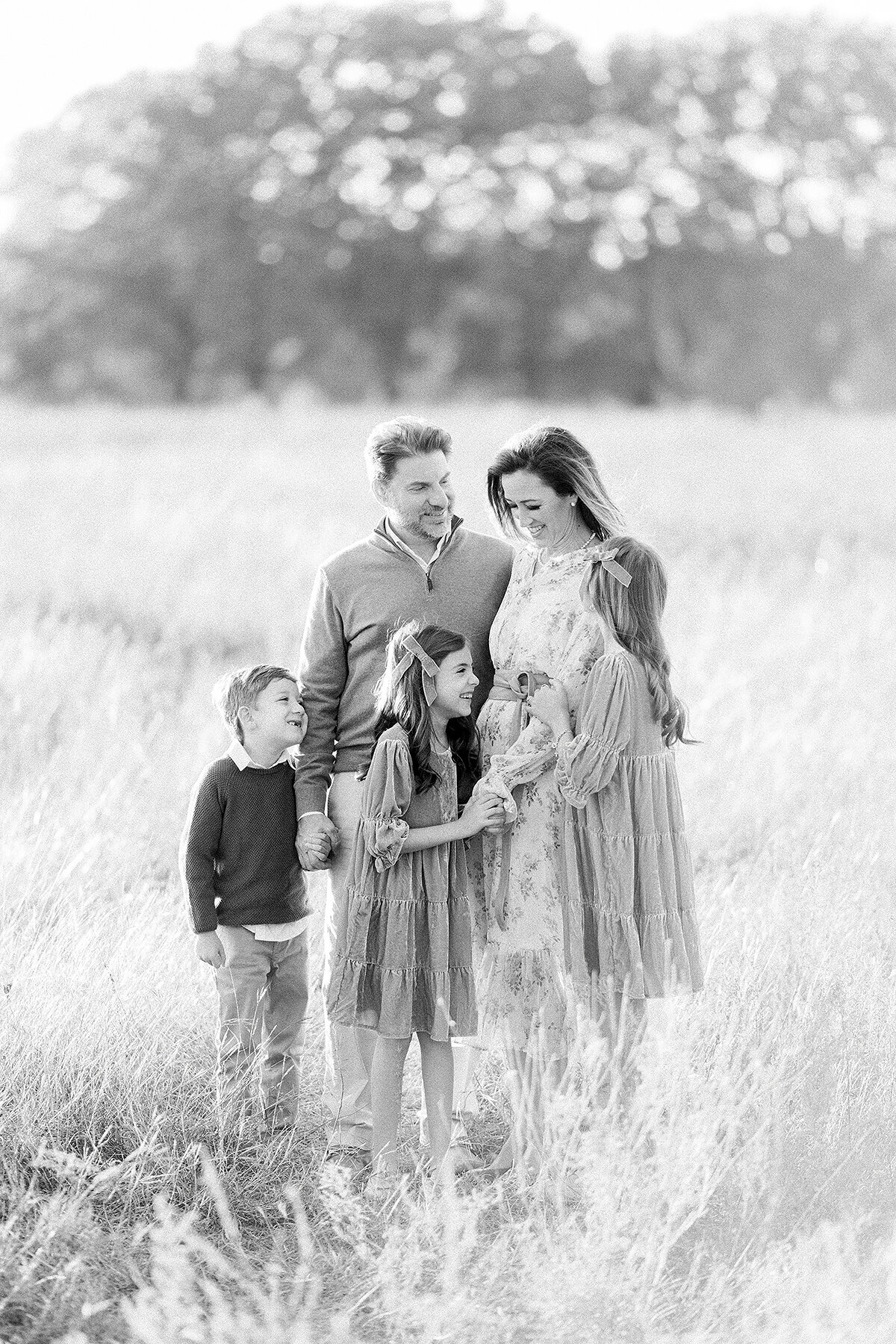 A black and white photo of a family of 5 taken while they are standing together holding each other in an open field at a local Dallas Texas park as their Dallas Family Photographer captures them together.