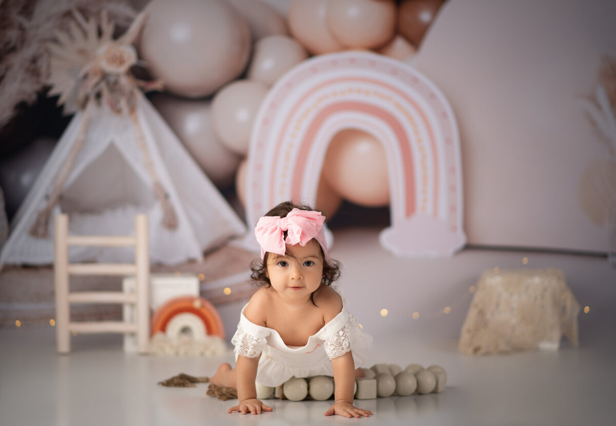 one year old baby girl wearing pink bow crawling in front of boho birthday backdrop