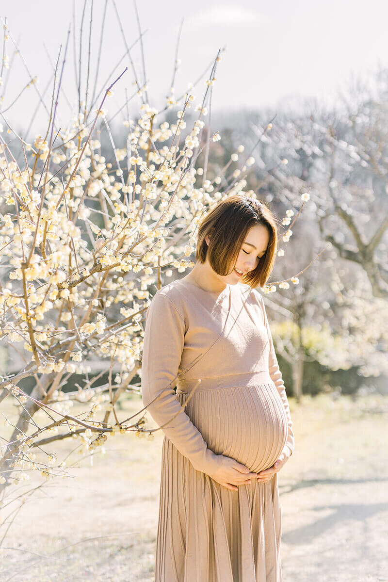 冬の2月、蠟梅が咲いている場所で、おなかに手を当ててたたずんでいる妊婦さんの美しいおしゃれなマタニティフォト。