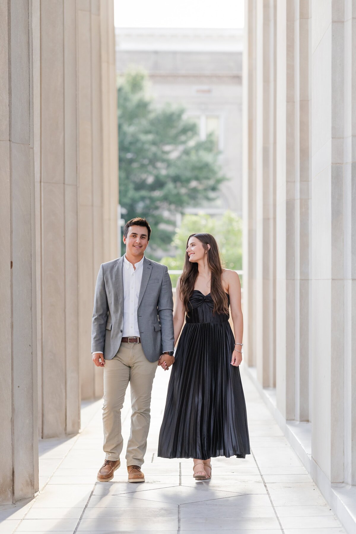 A woman in a black gown walks through a colonnade holding hands and smiling at her fiancee