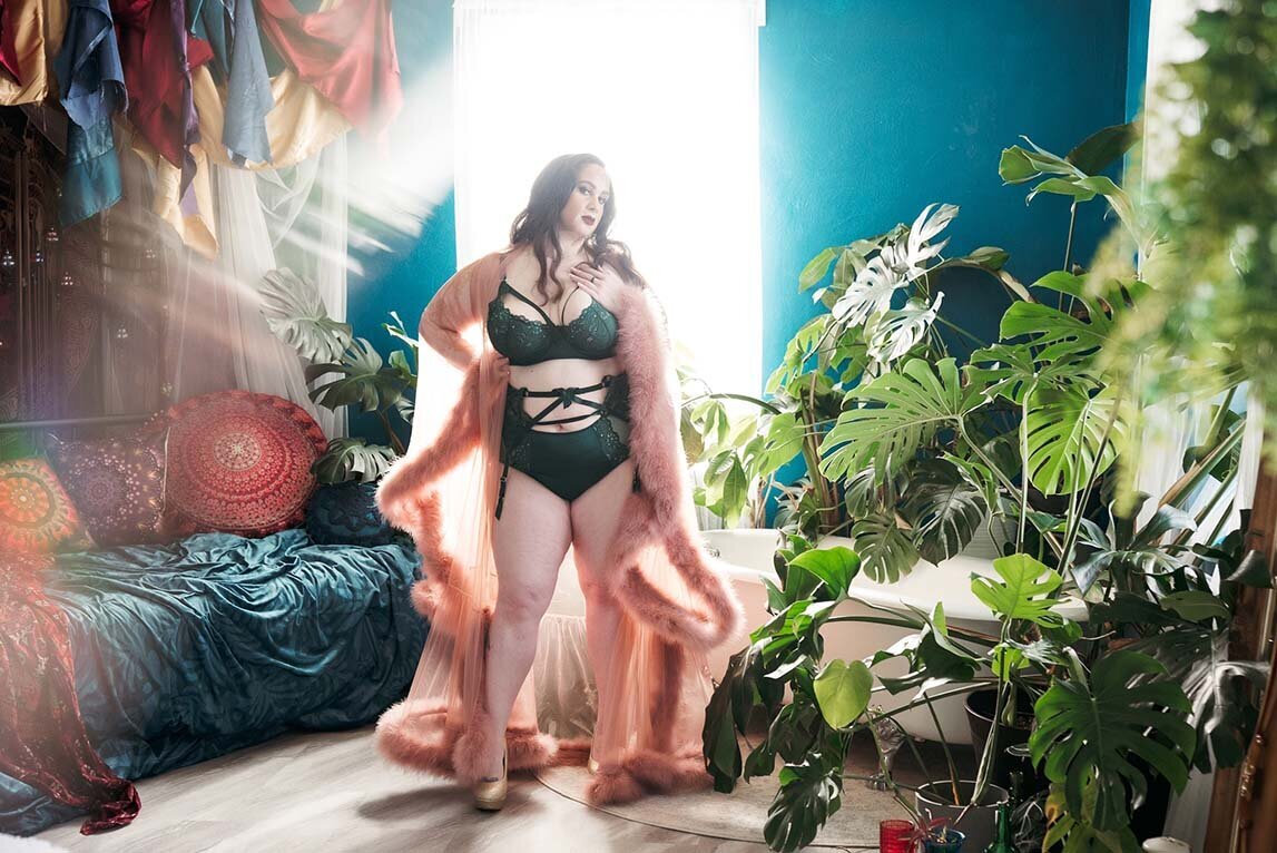 Boudoir portrait of curvy woman with red hair posed standing wearing a dark green lingerie set and pink robe in a bohemian styled boudoir set