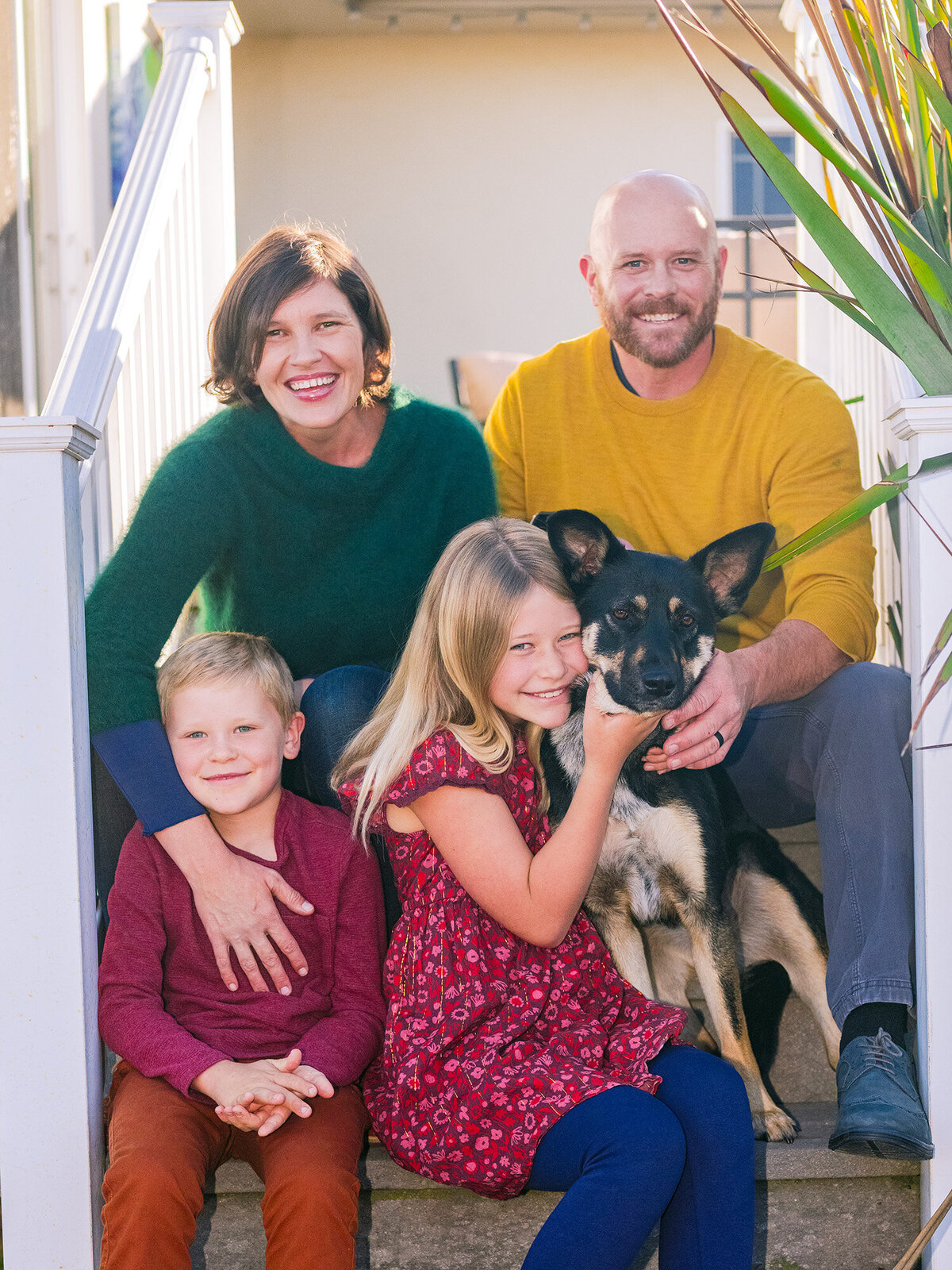 Smiling family with young children and their dog sitting on porch steps during a Marin County family photo session by Isela Lozano