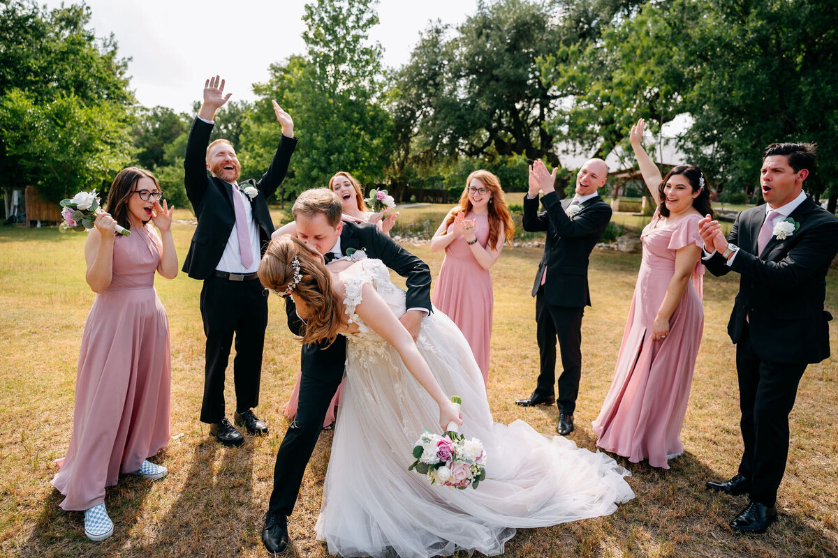 Groom dipping and kissing bride with cheering wedding party