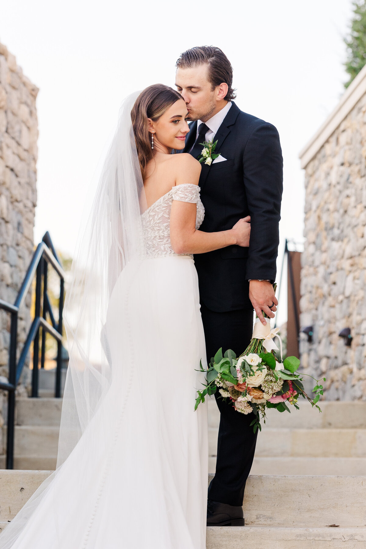 A romantic Westlake wedding moment with the groom kissing his bride on the forehead, framed by stone architecture and soft evening light near the Dallas-Fort Worth area.