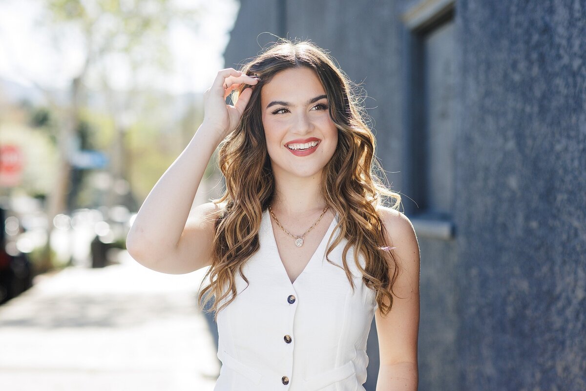 Sunlit senior girl smiling while walking in an urban Los Angeles alleyway wearing a chic white button-up romper for her modern senior portraits