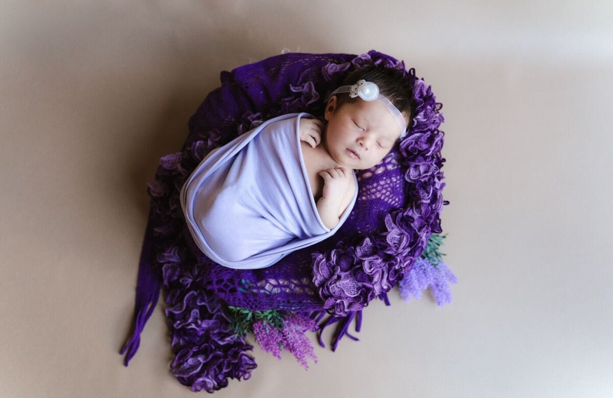 Baby in purple wrap newborn baby girl peacefully sleeping, swaddled in a soft lavender wrap, resting on a textured deep purple blanket with tiny lavender flowers.