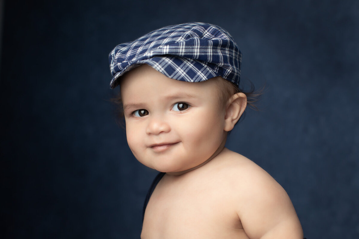 A baby with a mischievous smile poses confidently in a blue plaid newsboy cap against a deep blue backdrop. This classic portrait by one of the top baby photographers in Dallas captures personality and style in a timeless way.