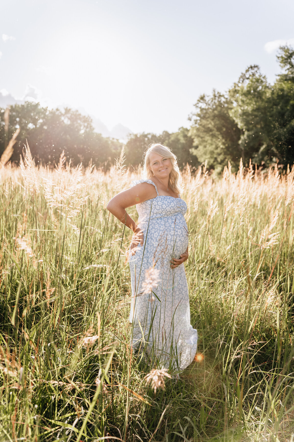 A pregnant woman stands confidently in a sunlit field, cradling her belly with one hand and resting the other on her hip, wearing a light blue floral dress.