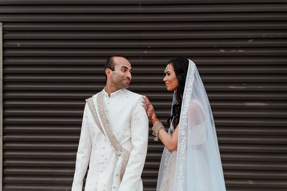 Hindu groom wearing a white sherwani with a light blue and gold dupatta looks over his shoulder and smiles at the Indian bride wearing a white beaded saree with a light blue dupatta veil. Her wedding bangles are white and gold and her hands are covered in wedding henna from her mendhi.
