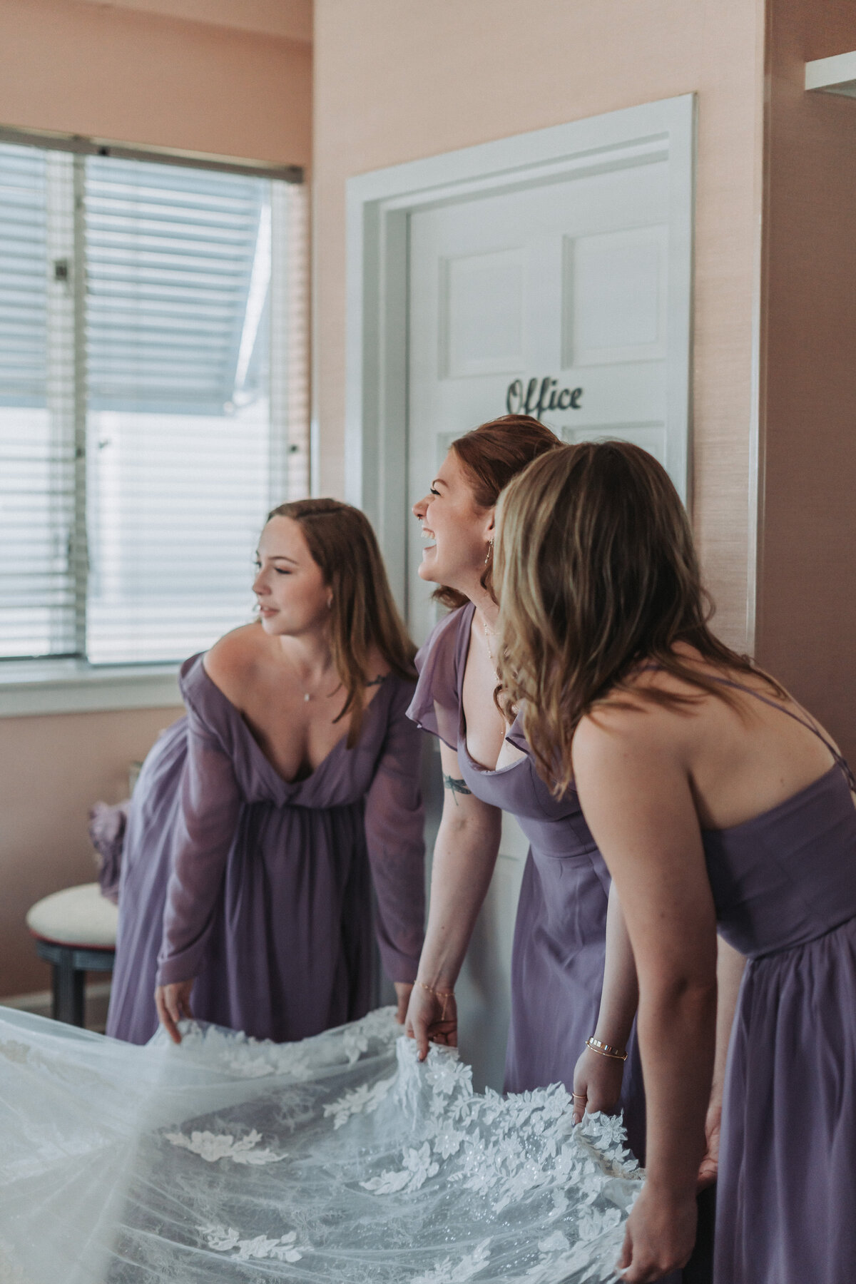 Bridesmaids in dusty lilac gowns lovingly adjust the intricate lace train of a bride's wedding dress, capturing a joyful pre-ceremony moment at the Lesner Inn in Virginia Beach, Virginia, by Quinn Photography LLC.