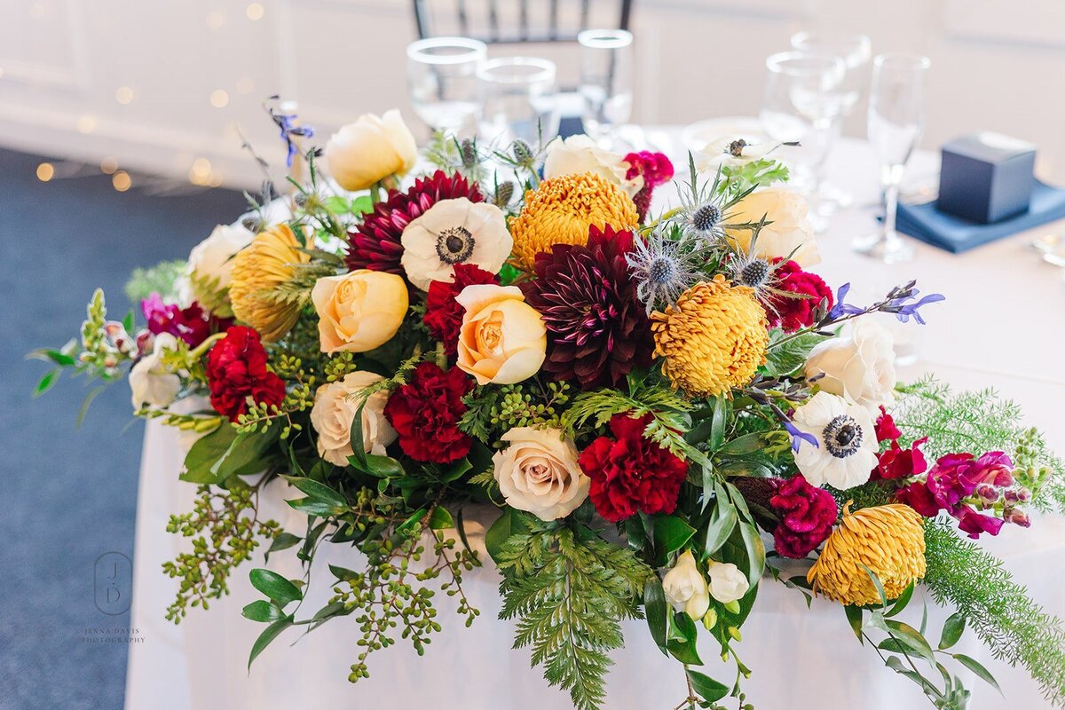 Artistic sweetheart table arrangement featuring fall colors and ferns. Creative floral design for weddings and special events at venues near Kent Island, MD and Tilghman Island, MD.