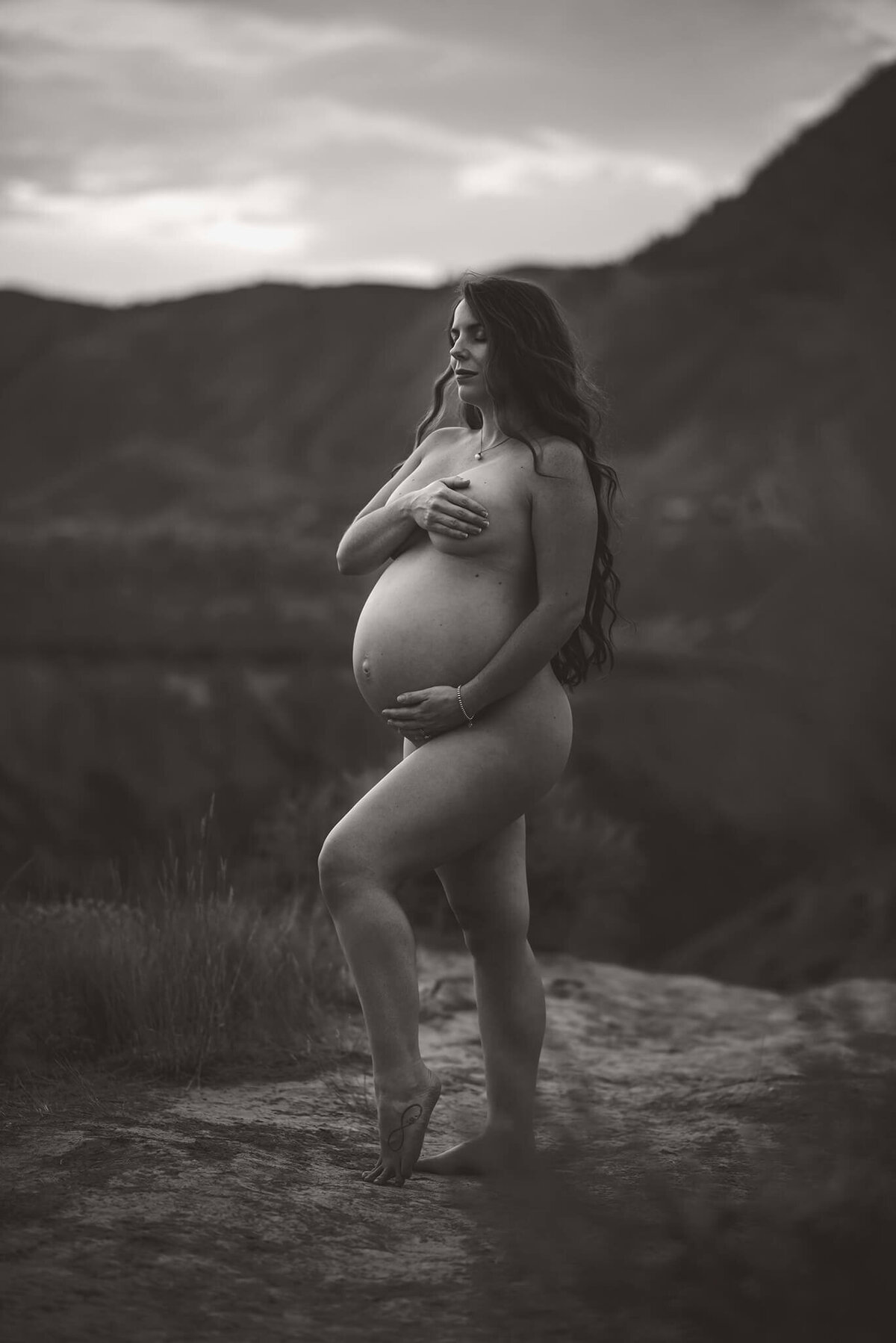 Brittany Danielle captures a stunning black and white portrait of a pregnant woman with long black hair. She stands nude covering her breasts and cradling her belly.