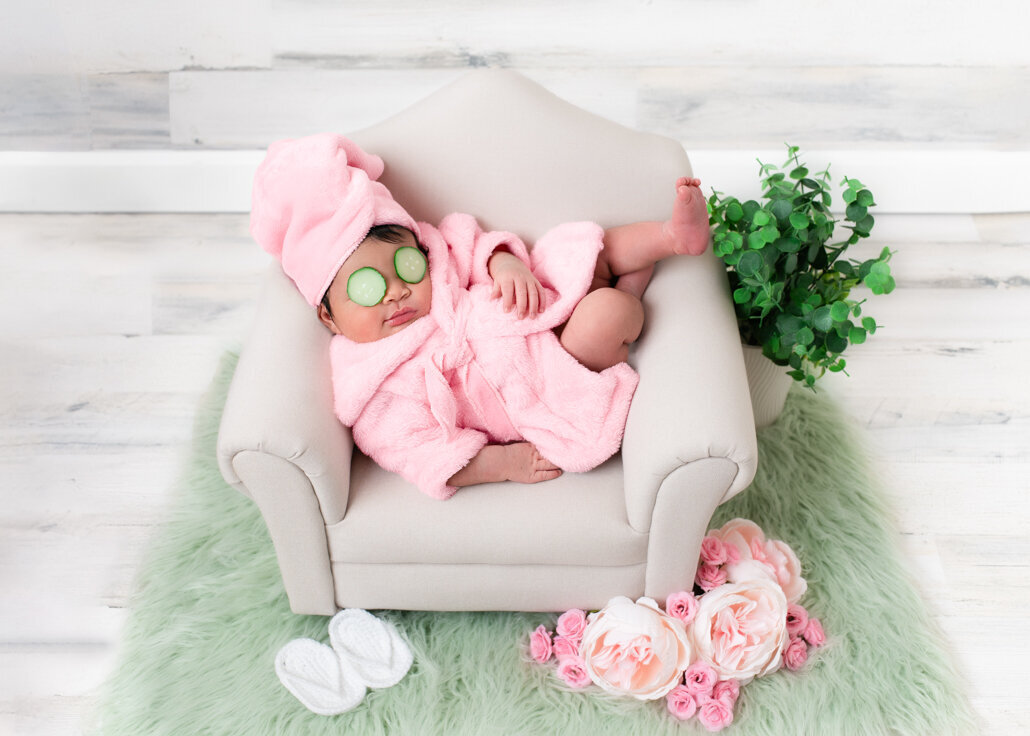 Newborn photography -- baby girl in a pink robe with cucumber eye pads, relaxing in a tiny chair with flowers and slippers in a Birmingham photography studio