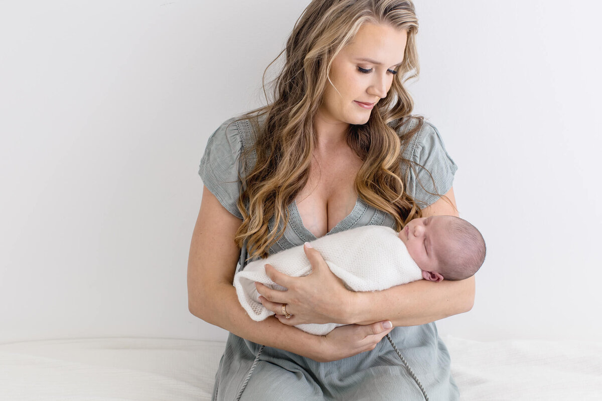 A woman with long hair gently cradles a sleeping baby wrapped in a white blanket. She is seated against a plain white background, gazing affectionately at the infant.