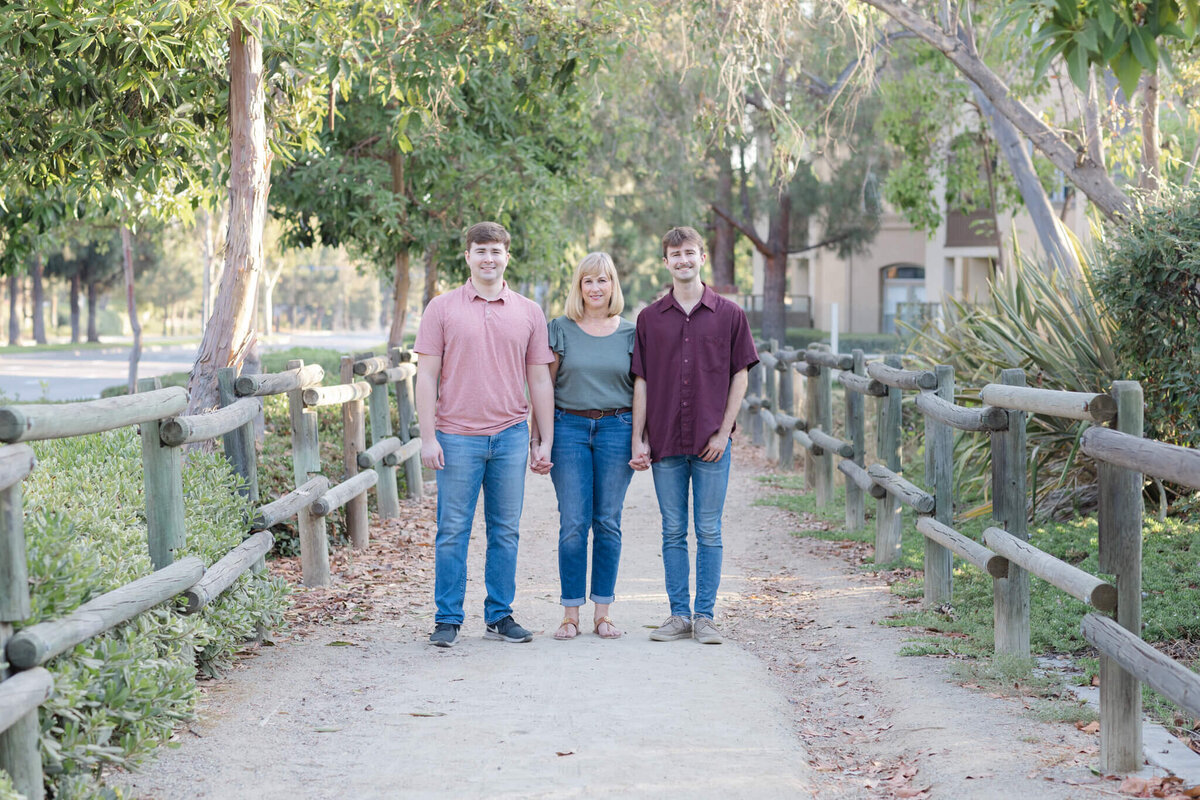 A mother with blonde hair in the middle of her two adult sons, holding their hands and standing facing the camera in a gravel pathway at Citrus Ranch Park in Tustin.