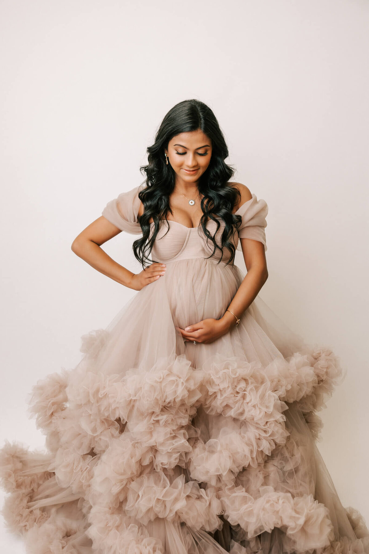 woman with long black hair standing in tan tulle gown smiling down at pregnant belly